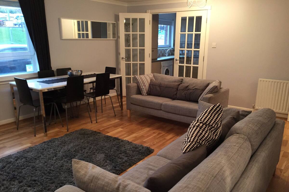 A living room with a grey sofa, a dining table with chairs, and a kitchen visible through an open door.