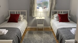 A bedroom with two single beds, a nightstand, a lamp, and a window with curtains. - Serviced Apartments East Kilbride (Glasgow)