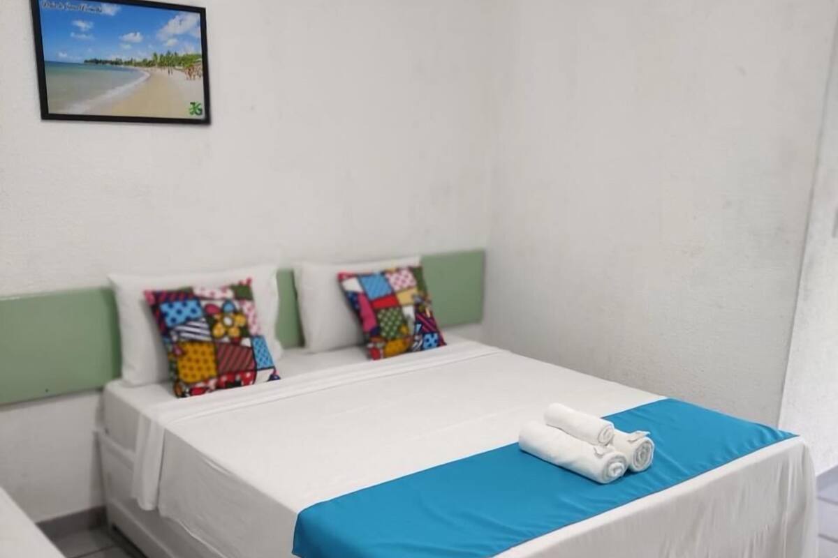 A single bed with a blue bedspread and two colorful pillows, a white headboard, and a framed picture of a beach on the wall.