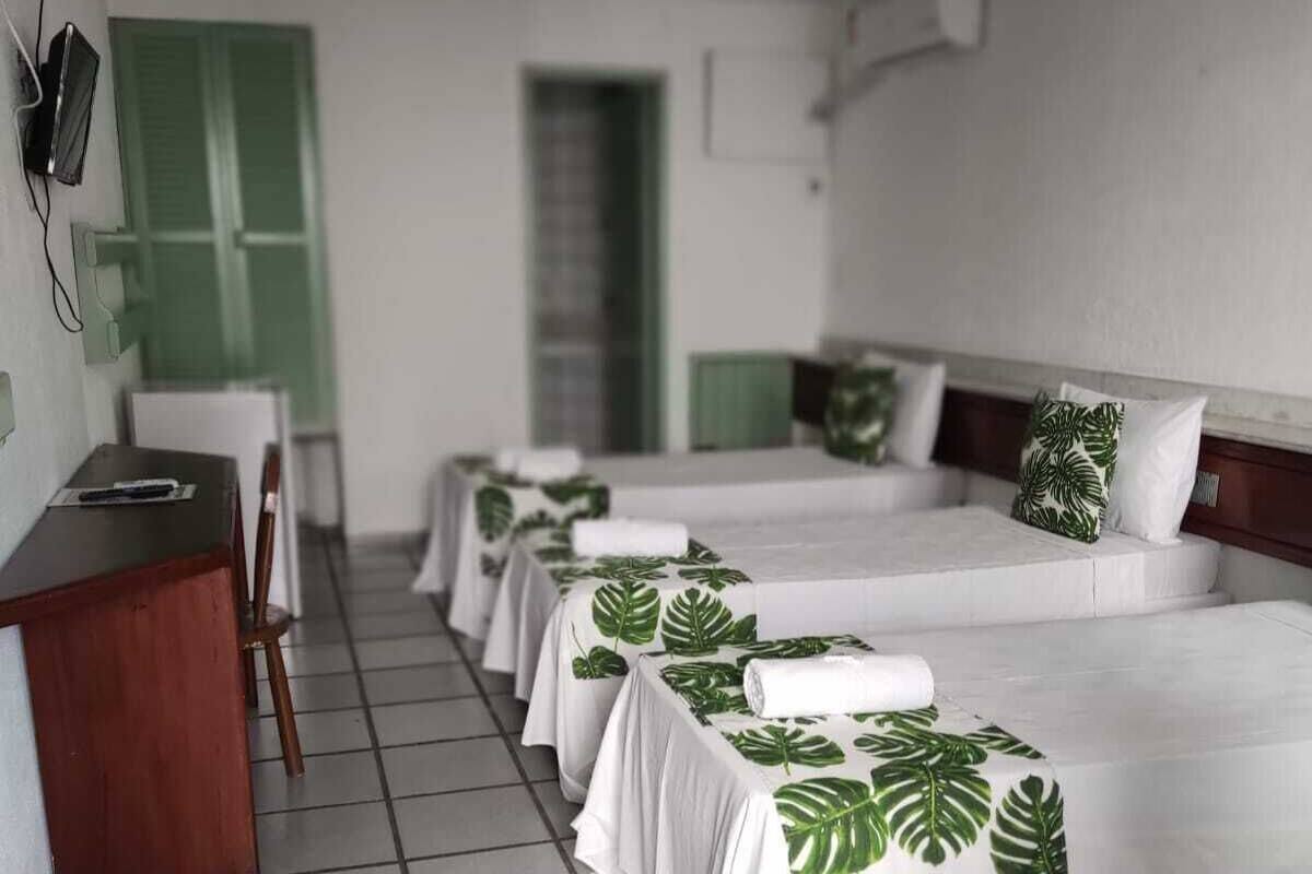 A hotel room with four beds, each with a white bedspread featuring green leaf patterns, a wooden desk, and a chair.