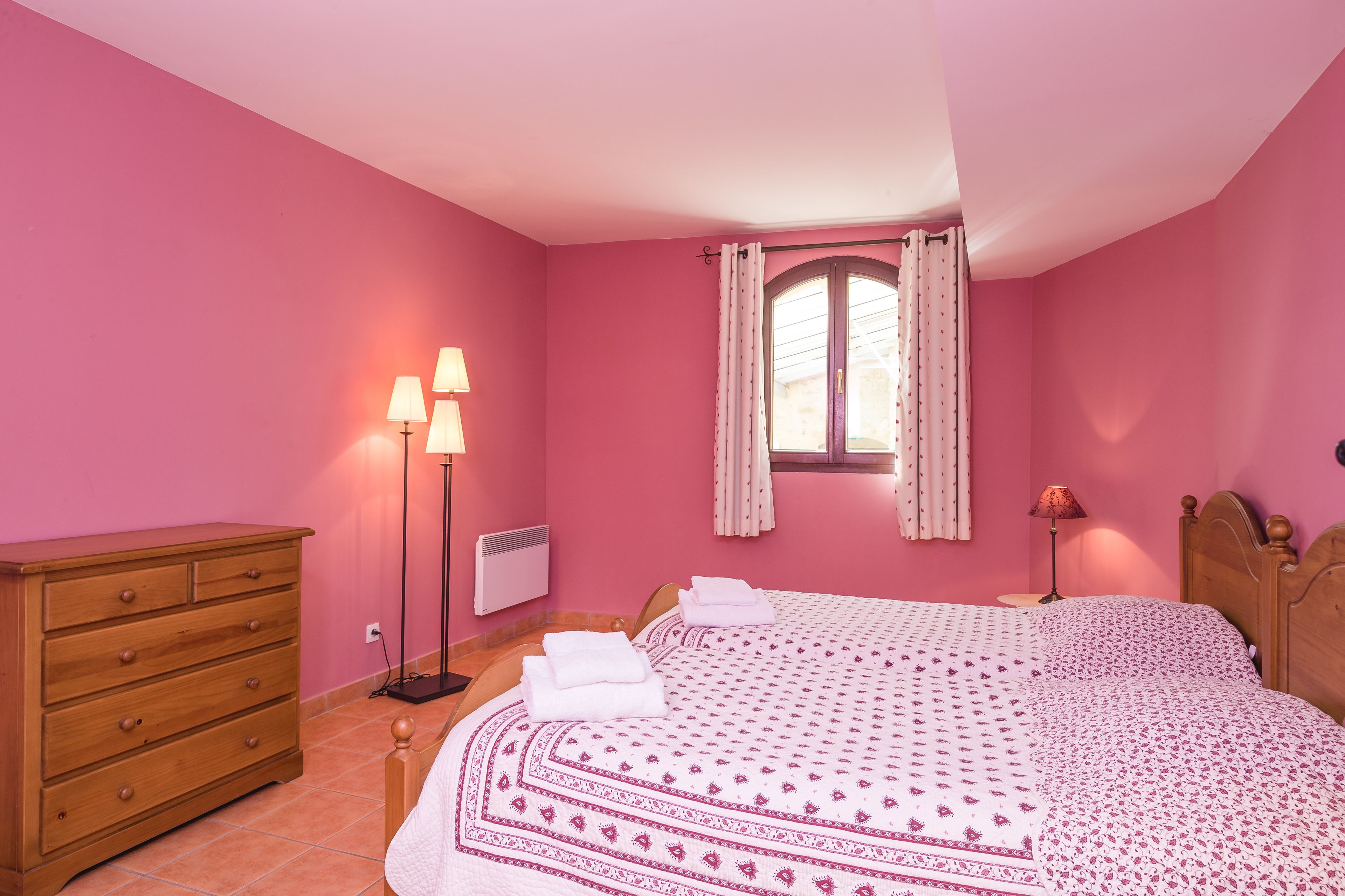 Une chambre avec des murs roses, une commode en bois, un lit avec une literie à motifs, deux lampes de chevet et une fenêtre avec des rideaux.