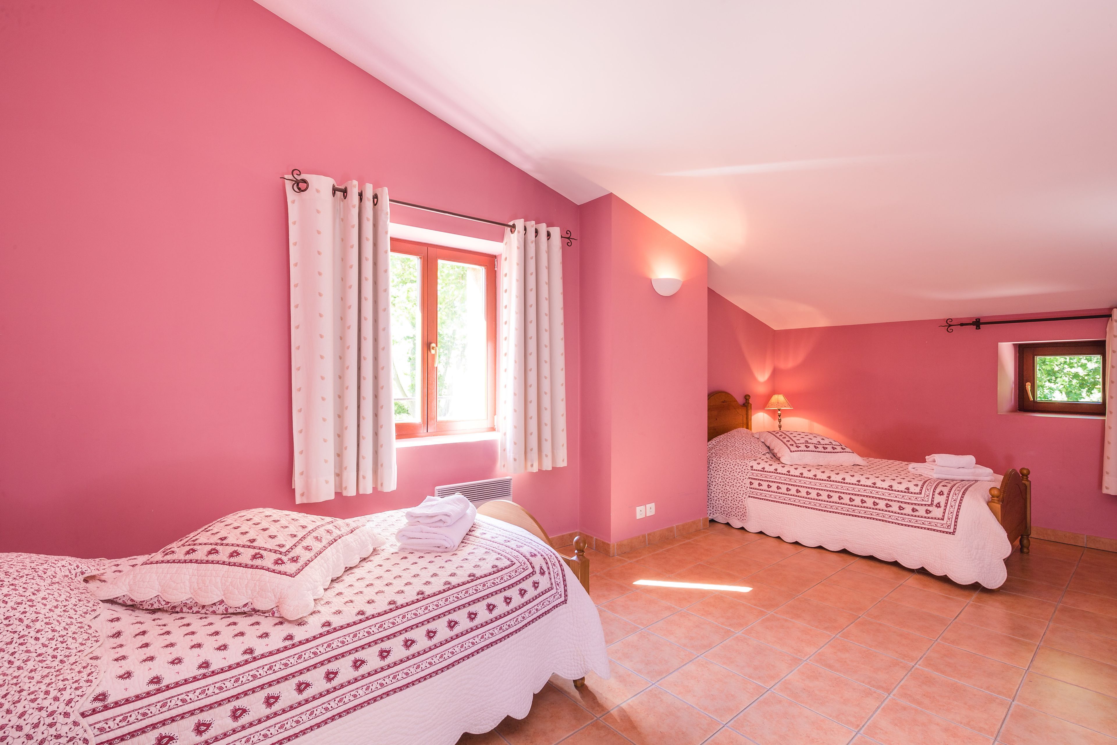 A pink-themed bedroom with two beds, a window with curtains, a wall-mounted lamp, and a tiled floor.