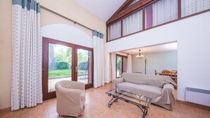 A spacious living room with a sofa, armchair, and glass coffee table. A large sliding glass door leads to a garden. - Le Relais du Grand Logis Cadarache ITER (Mirabeau)
