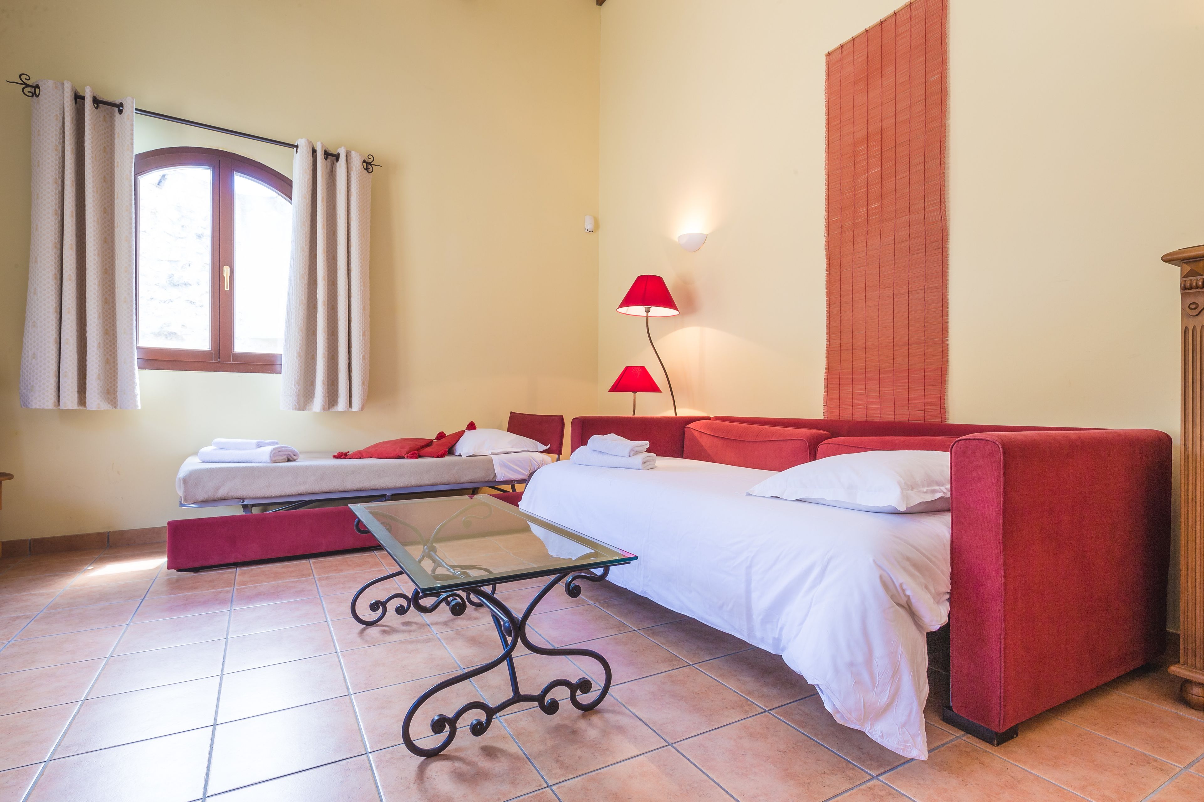 Une pièce comprenant un lit, un canapé rouge, une table en verre, une fenêtre avec des rideaux et une lampe fixée au mur.
