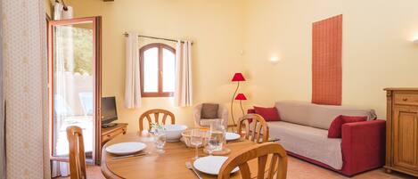Un comedor con mesa de madera, sillas y un sofá. Hay un televisor sobre un mueble bajo, una lámpara y una ventana con cortinas.
