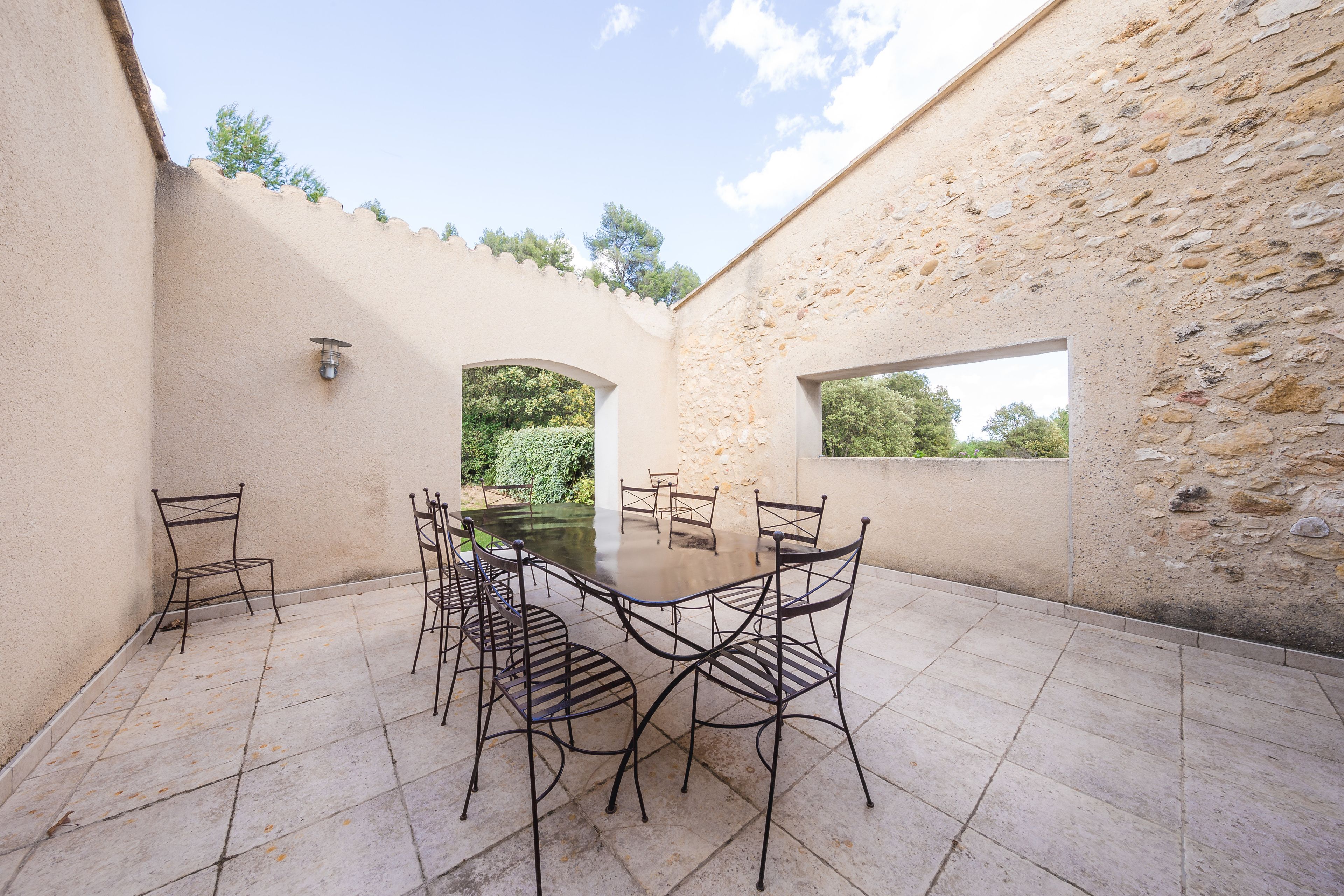 Un patio exterior con una mesa redonda y sillas de metal, rodeado de muros de piedra y vistas al verdor.