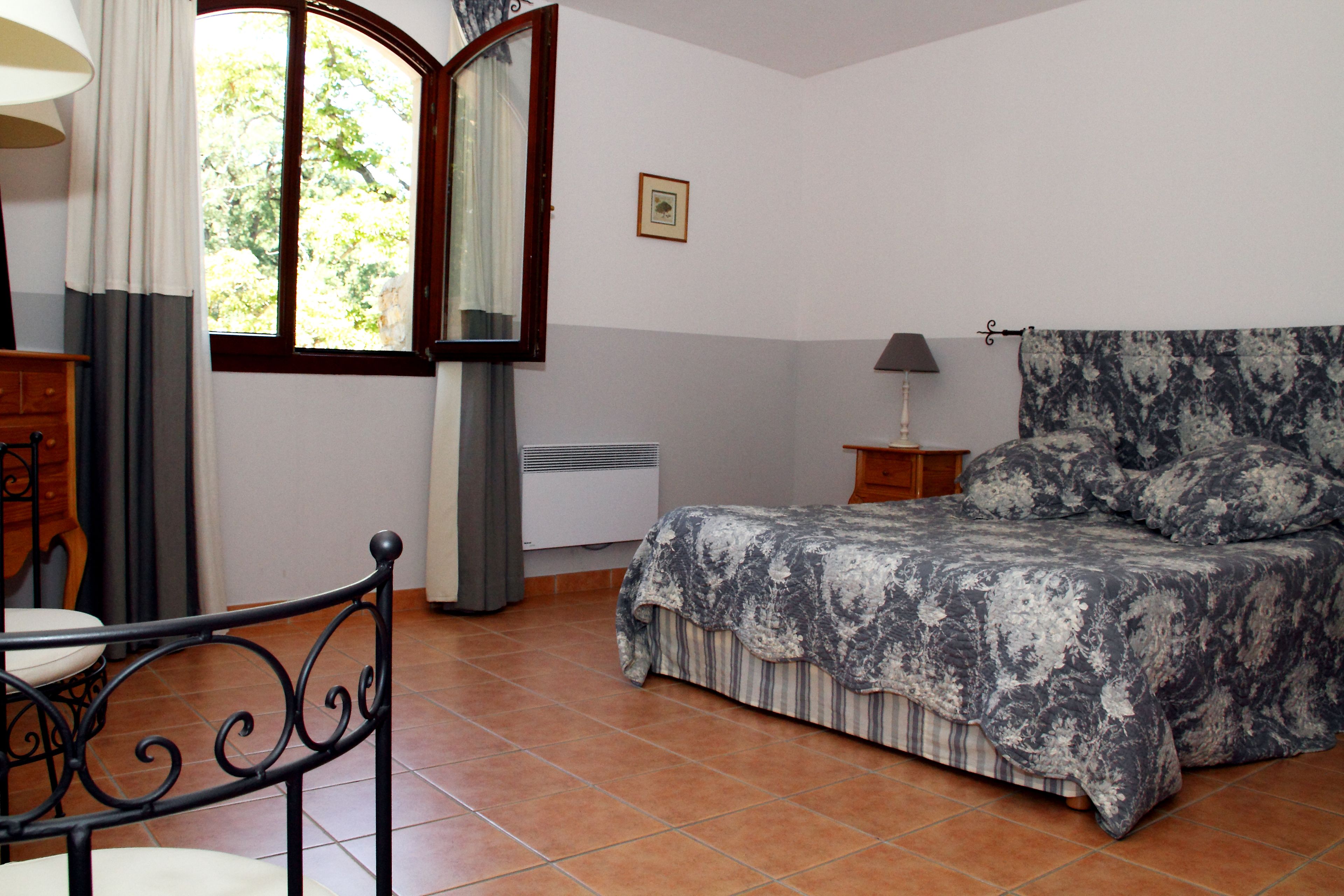 A bedroom with a bed, a chair, a nightstand, a lamp, and a window with curtains.