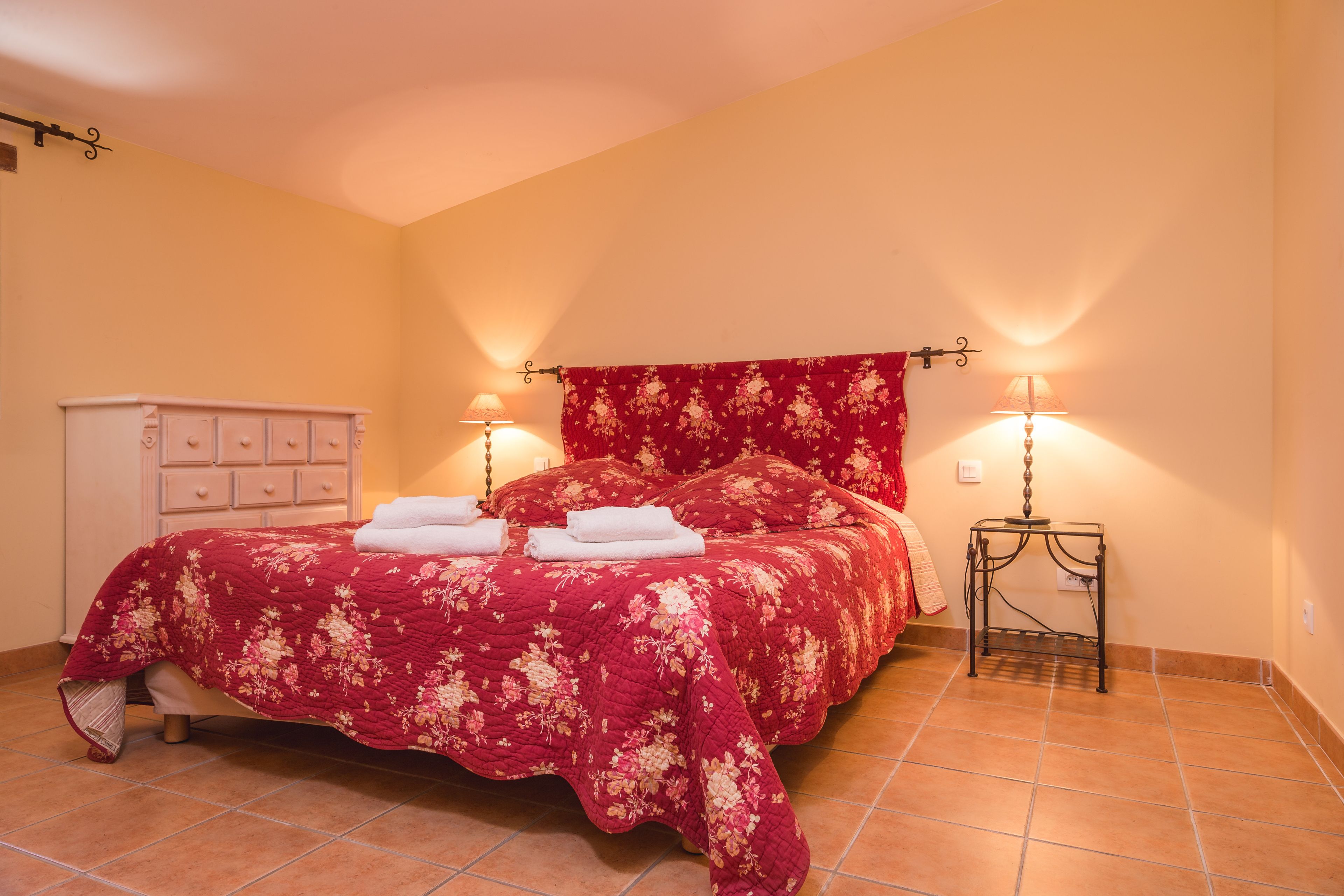 Un dormitorio con una cama, dos lámparas de noche, un tocador blanco y una colcha roja con estampado floral.