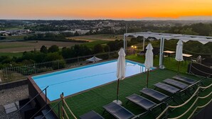 A swimming pool with lounge chairs and umbrellas, surrounded by a fence and a view of the countryside at sunset.