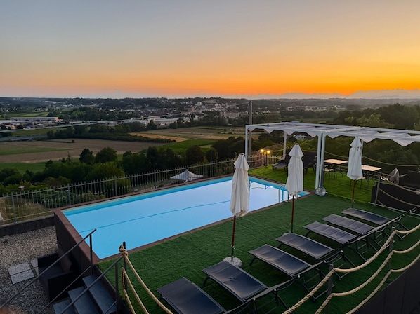 Une piscine entourée de chaises longues et de parasols, délimitée par une clôture, et offrant une vue sur la campagne au coucher du soleil.