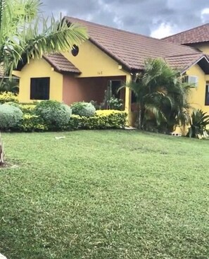 Uma casa amarela com telhado vermelho, rodeada por um gramado bem cuidado e diversas plantas.