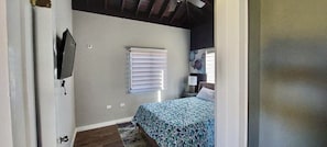 A bedroom with a bed, a window with blinds, a wall-mounted TV, and a door.