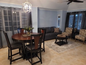 A living room with a round wooden dining table, dark chairs, a sofa, patterned armchairs, a coffee table, and a ceiling fan.