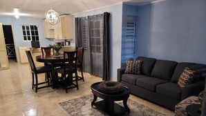 Una sala de estar con sofá, reposapiés estampado y una mesa de centro con un adorno decorativo. Se ve un área de comedor con mesa y sillas al fondo.