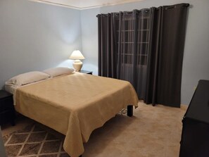 Quarto com cama, mesa de cabeceira, luminária e janela com cortinas.