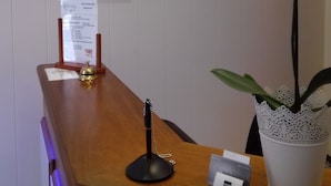 Un mostrador de recepción con detalles de madera, un portalápices negro, una flor blanca en una maceta blanca, y dos lámparas fijadas a la pared.