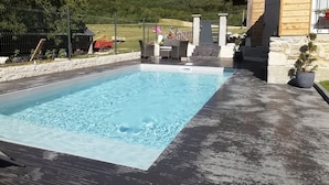 Una piscina rectangular con una cubierta de madera alrededor, paredes de piedra y vista a una colina boscosa al fondo.