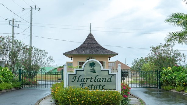 Une entrée fermée par un portail, sur laquelle on peut lire l’inscription « Hartland Estate », le tout entouré d’une végétation luxuriante et agrémenté d’un petit bâtiment doté d’un toit en pente.
