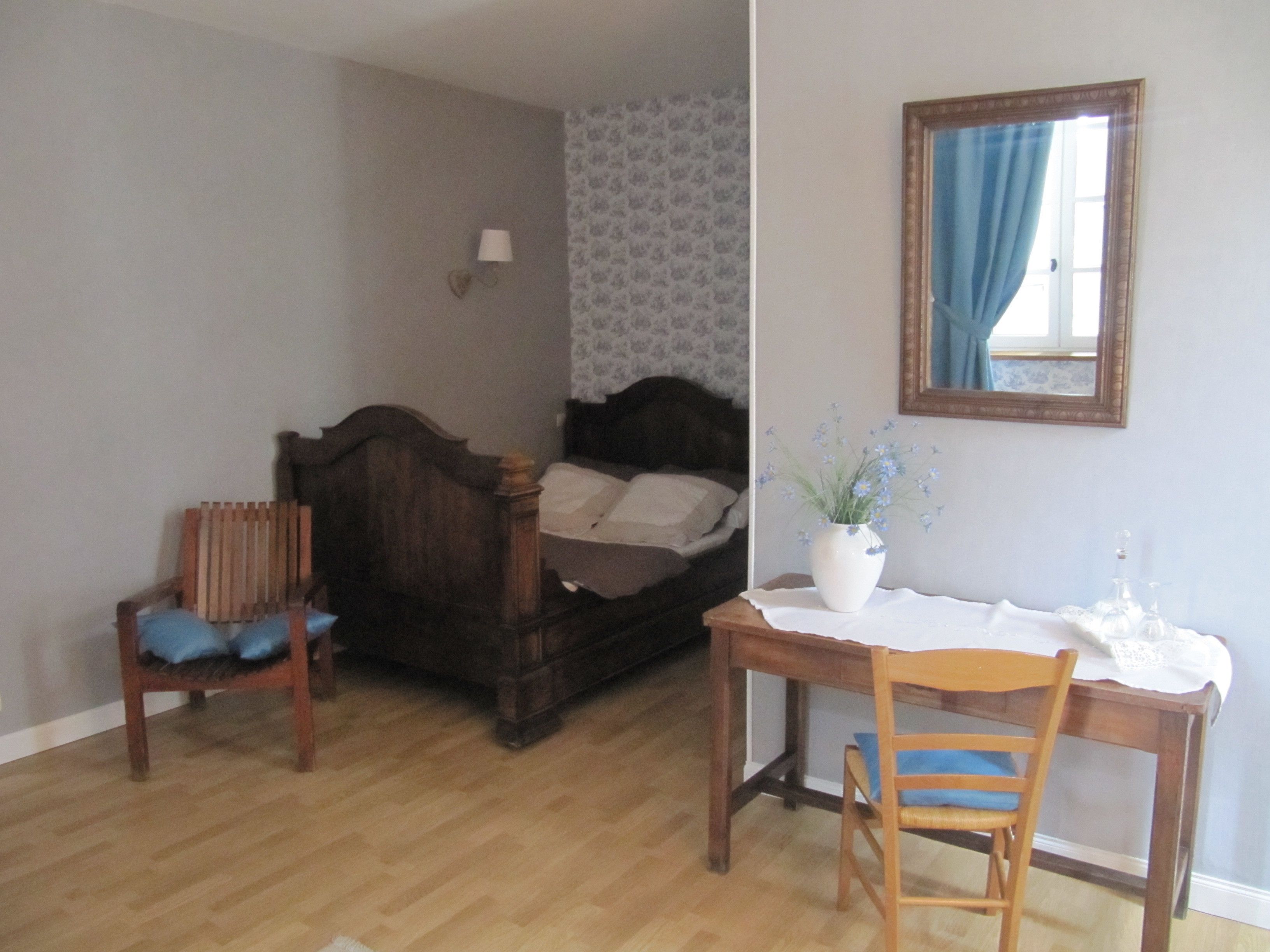 A bedroom with a wooden bed, a chair, a desk, and a vase with flowers.