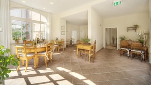 Uma sala de jantar ampla, com várias mesas e cadeiras de madeira, janelas grandes com cortinas e plantas em vasos.