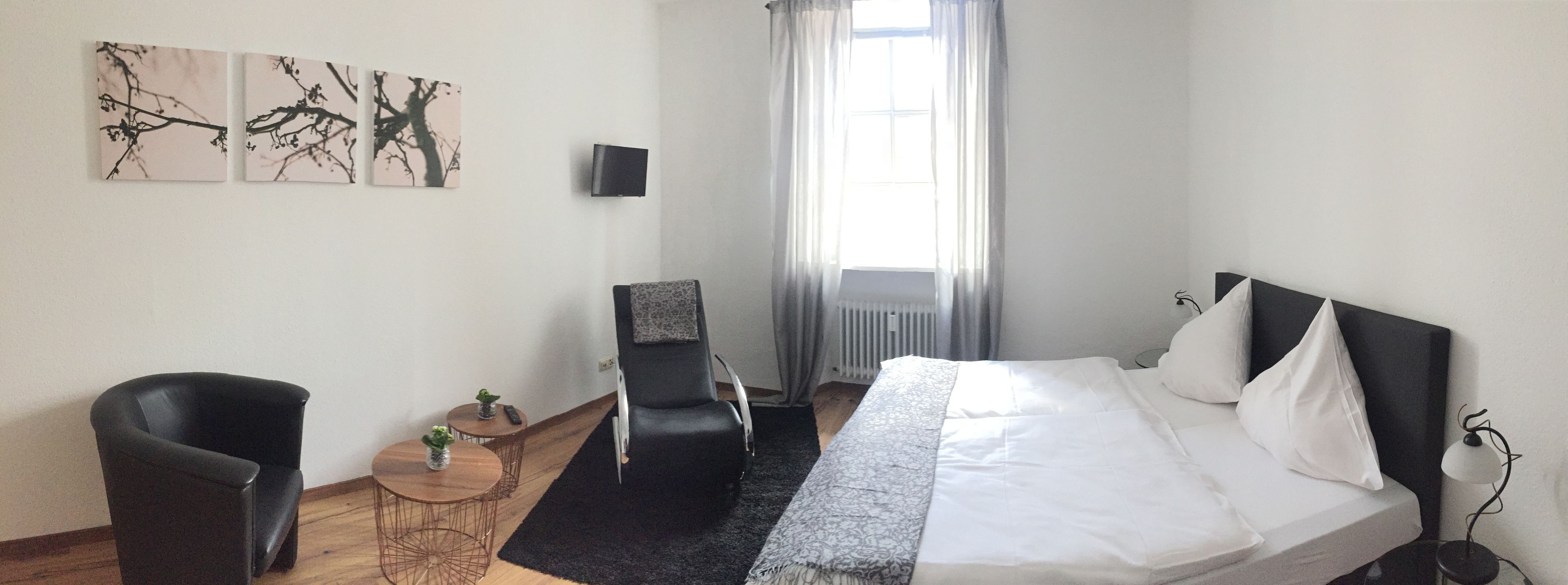 Habitación de hotel con una cama, dos sillas, una mesita con un jarrón y tres cuadros colgados en la pared.