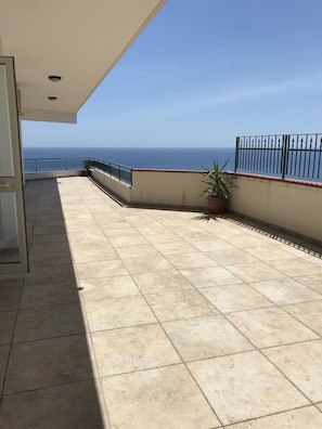 Un balcón con baldosas y vistas al mar, una planta en maceta y una barandilla de metal.
