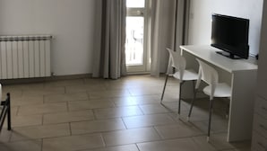 A room with a white desk, a television, and a chair. There is a radiator on the left and a window with curtains. - La nave sul mare di Taormina B&B (Letojanni)