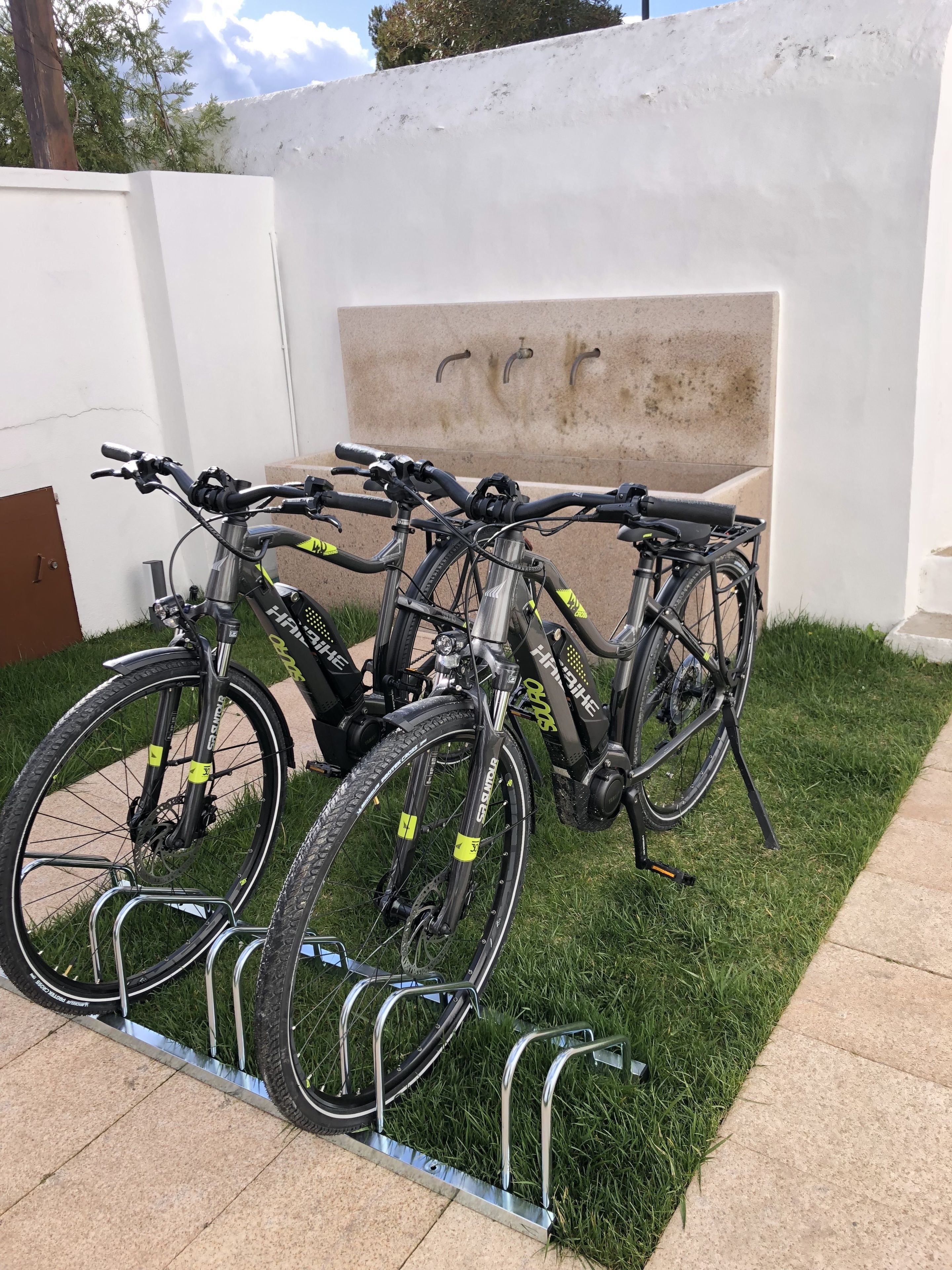 Varias bicicletas estacionadas en un césped, con un muro blanco y un banco al fondo.