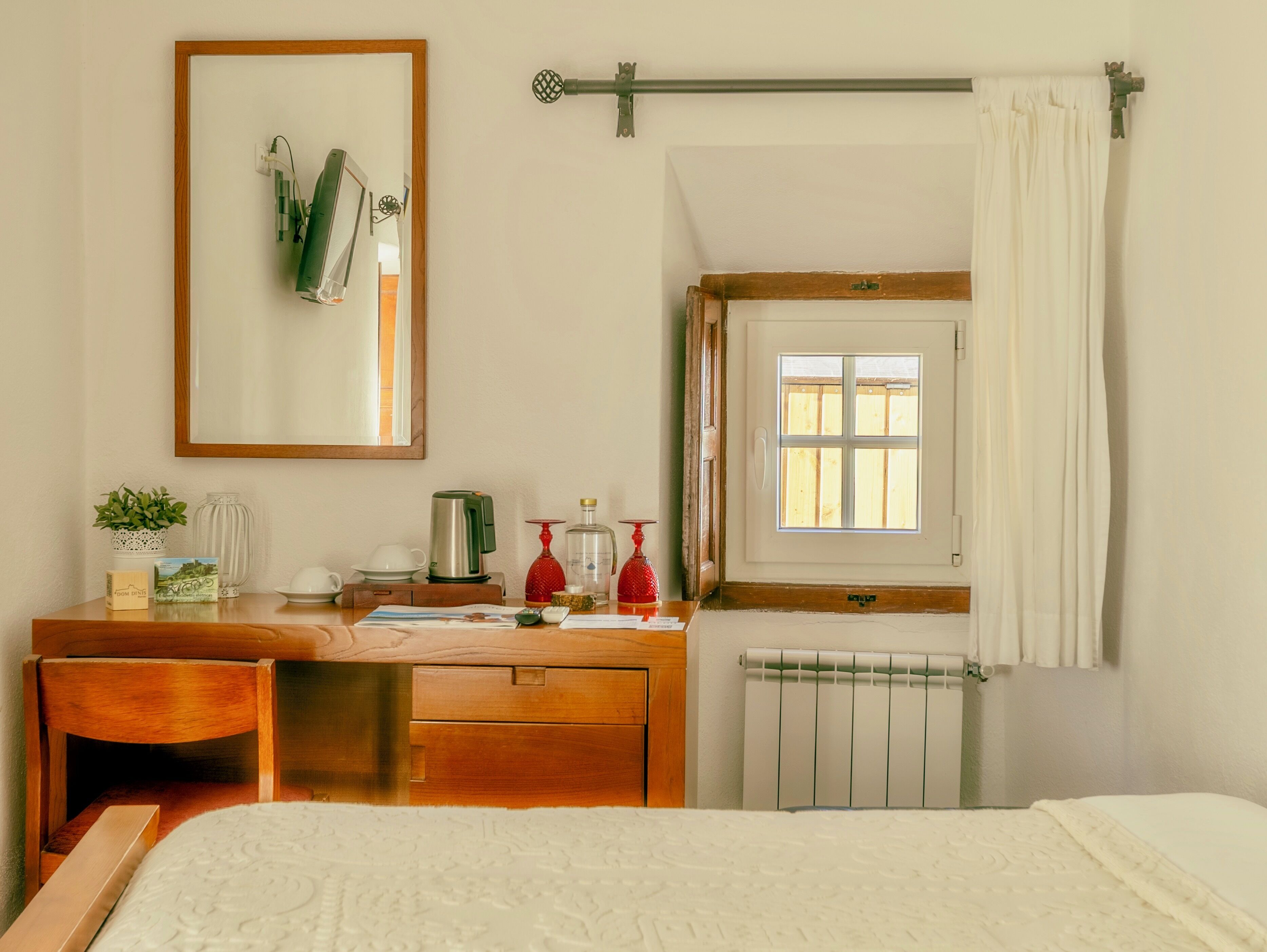 Ein Schlafzimmer mit einem hölzernen Schminktisch, einem Nachttisch, einem Heizkörper, einem Fenster mit Vorhängen und einem gerahmten Bild an der Wand.