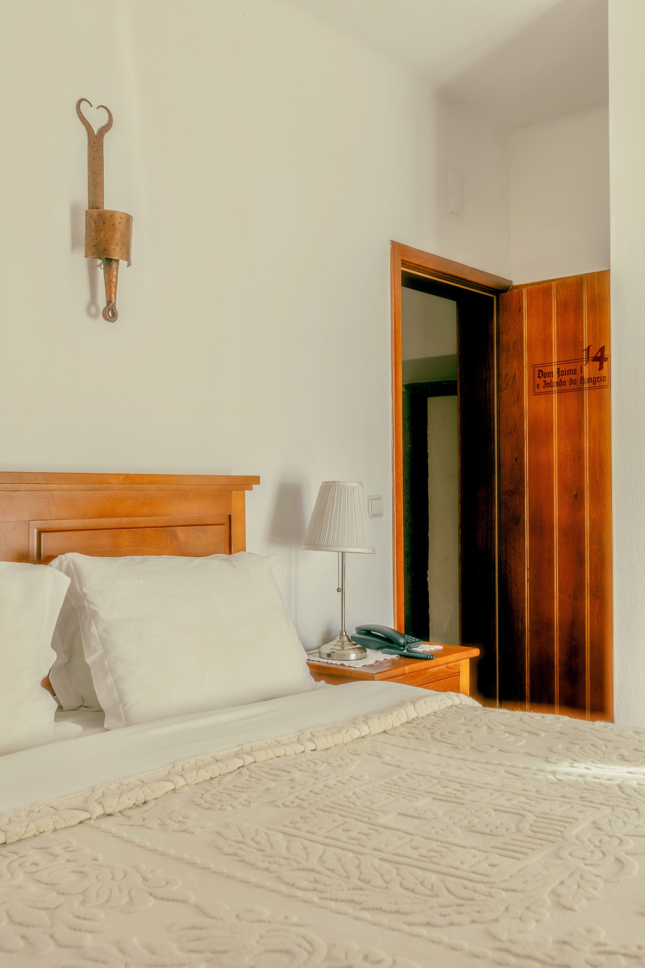 A bedroom with a wooden headboard, a bed with white bedding, a bedside lamp, and a wooden door with a number.