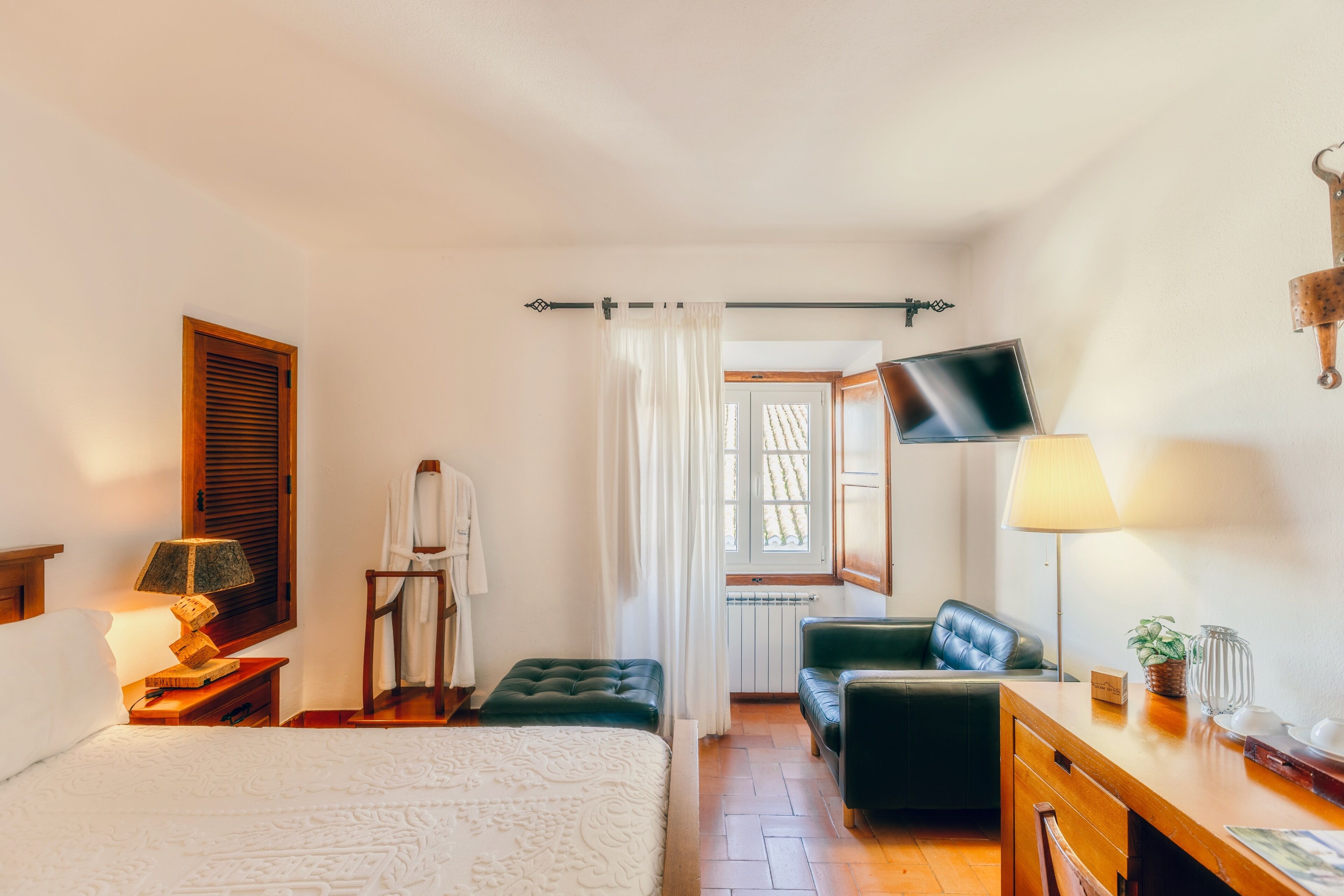 Habitación de hotel con cama, escritorio, silla, televisor, lámpara y ventana con cortinas.