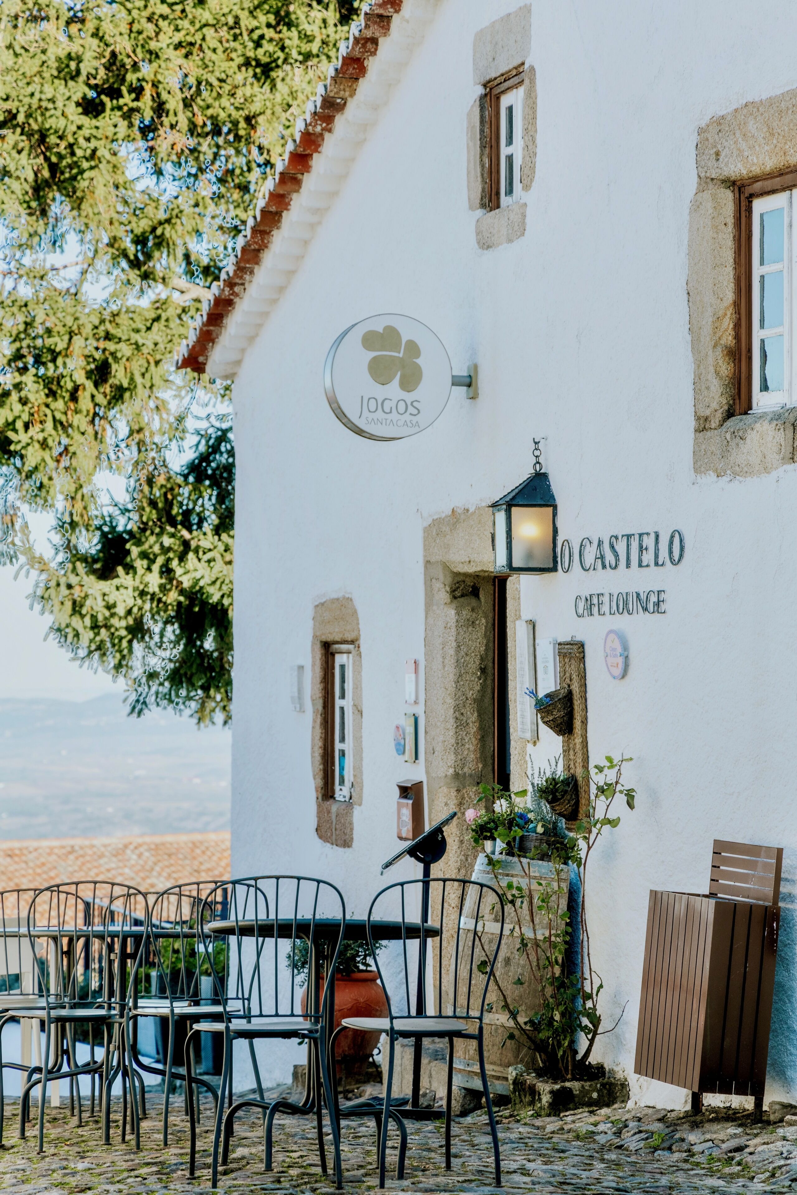 A building with a sign reading 'O CASTELO CAFE LOUNGE' and a logo above it. There are outdoor tables and chairs arranged in front.