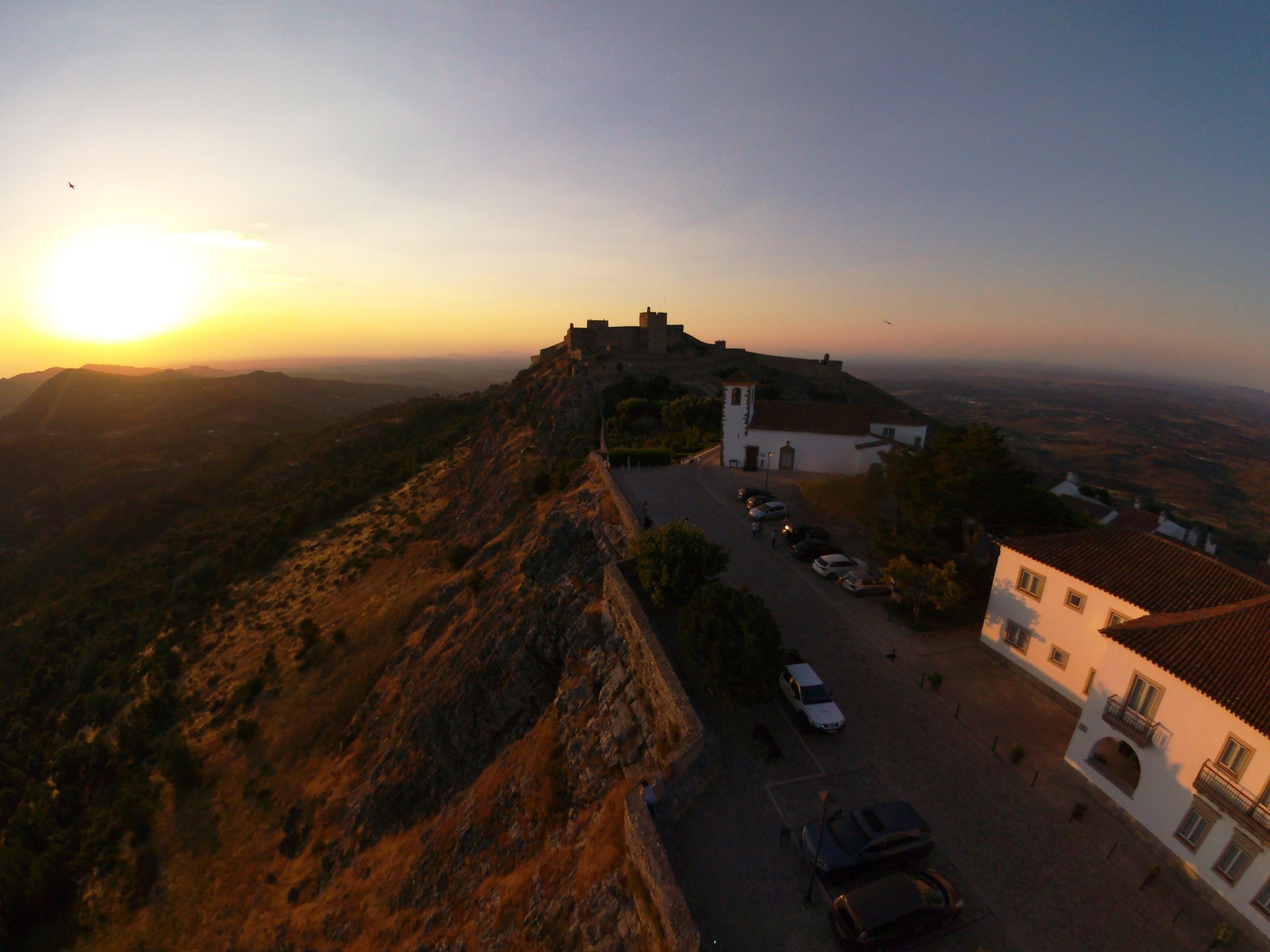 A fortified hilltop structure with a road leading to a white building with balconies.