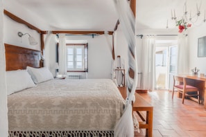 A bedroom with a large bed, wooden headboard, and a canopy. There is a desk and chair in the room, and a view of the outdoors through a window. - Dom Dinis Marvão (Marvao)
