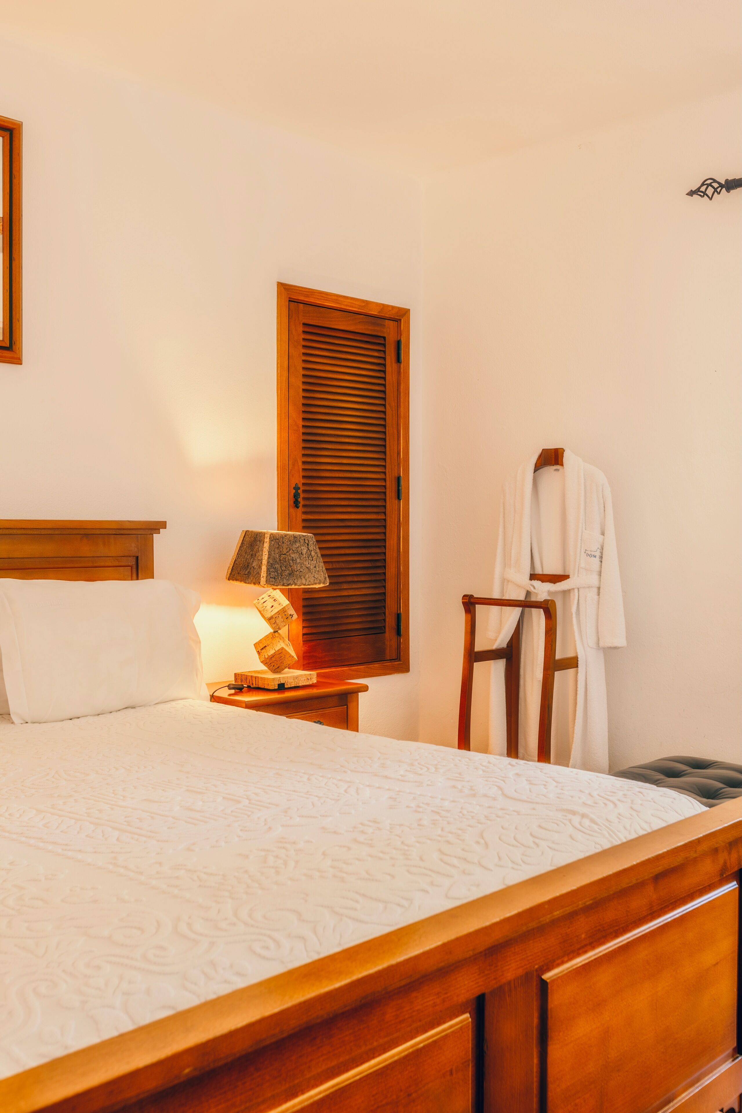 Een net opgemaakt bed met een houten hoofdbord, een bedlampje, een nachtkastje van hout, een houten stoel, en een witte badjas die aan een houten kapstok hangt.