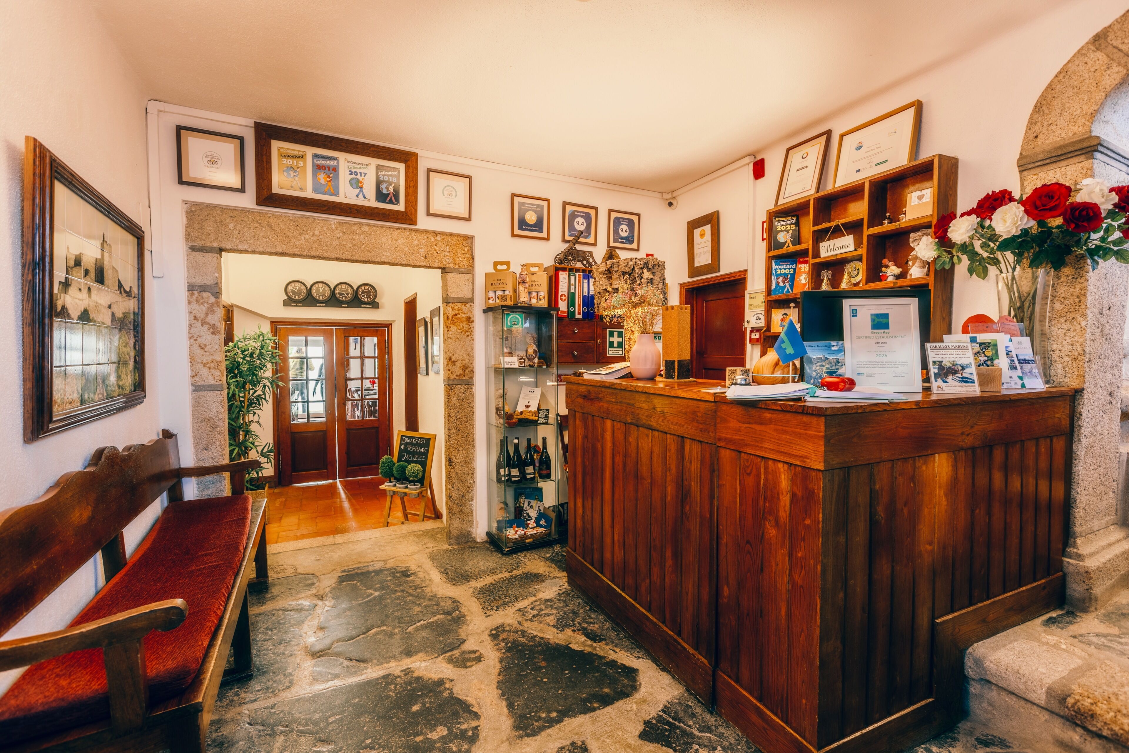 A reception area with a wooden counter, framed certificates, a welcome sign, and a vase with red and white flowers.