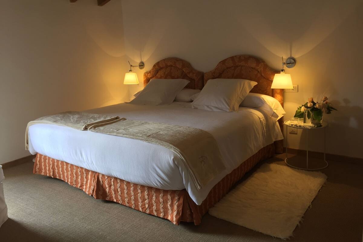 Ein ordentlich bezogenes Bett mit weißer Decke und orangefarbenem Bettrock, zwei Nachttische und ein kleiner Tisch mit einem Blumenstrauß.
