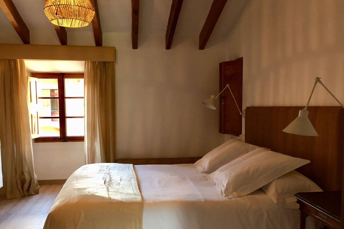 Ein Schlafzimmer mit Bett, Holzbalken, einer Hängeleuchte, einem Nachttisch und einem Fenster mit Vorhängen.
