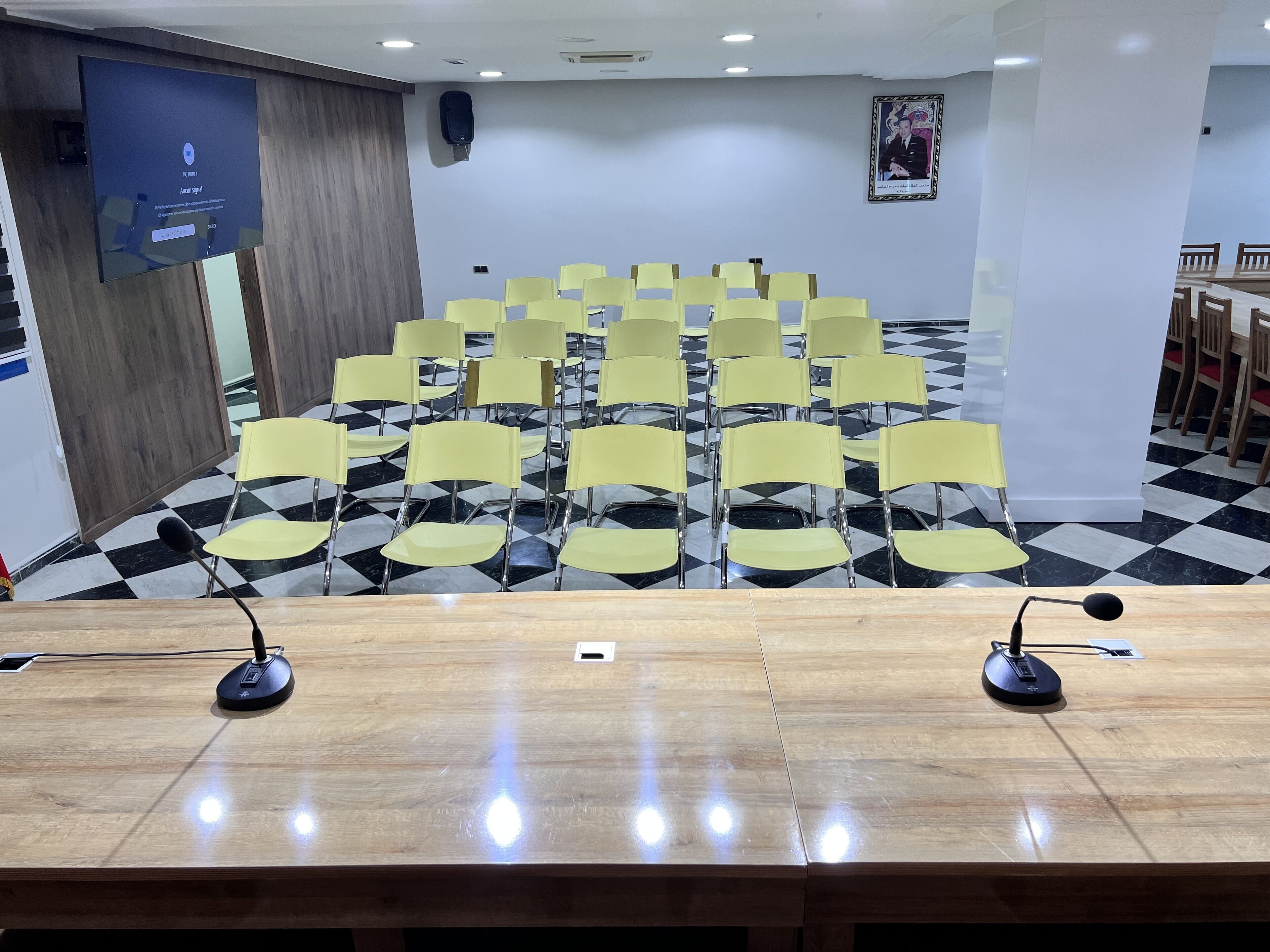 Un salón de conferencias con piso a cuadros, sillas amarillas, una mesa de madera con micrófonos y un altavoz en la pared.