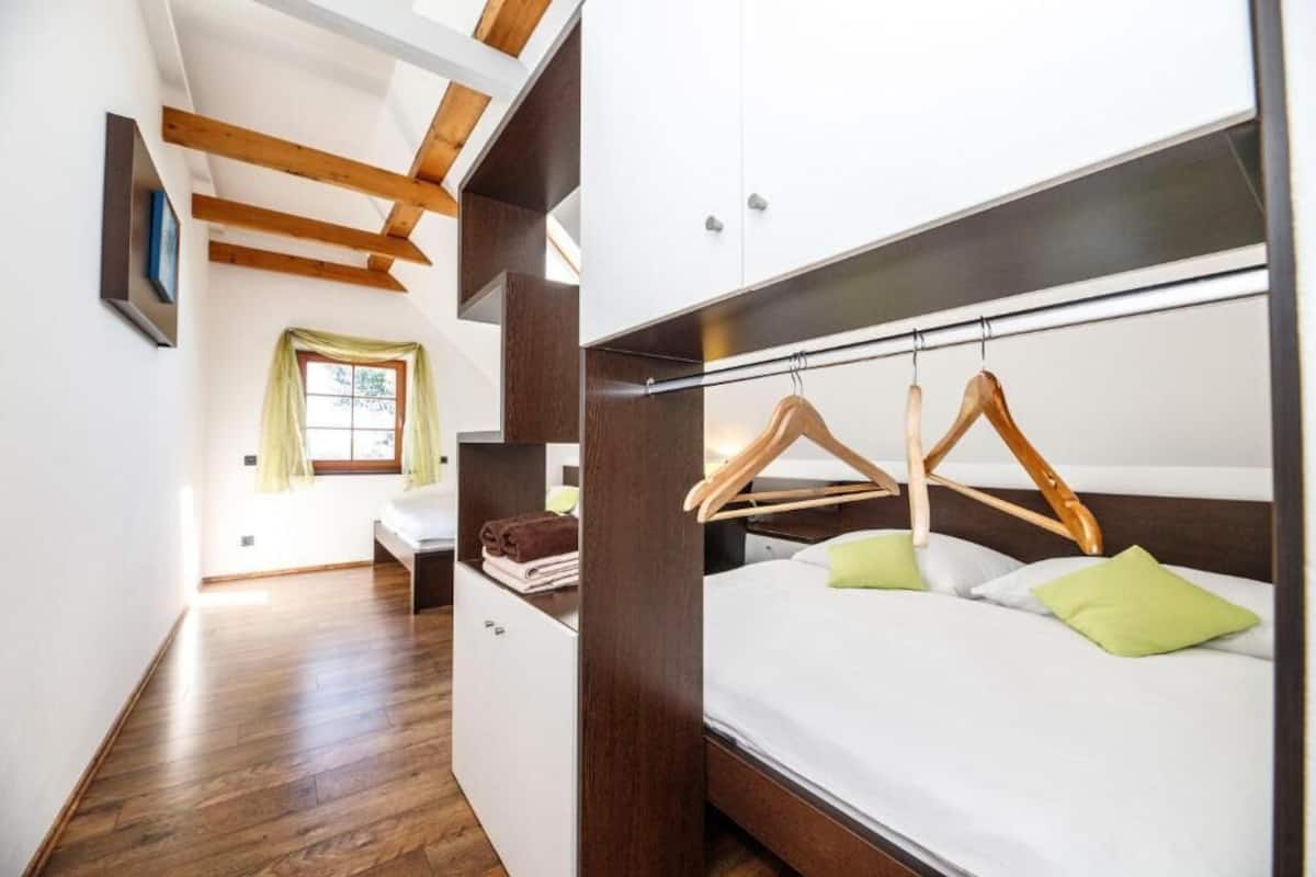 Um quarto com piso de madeira, uma cama com colchão branco e travesseiros verdes, um armário de madeira e uma janela com cortina.