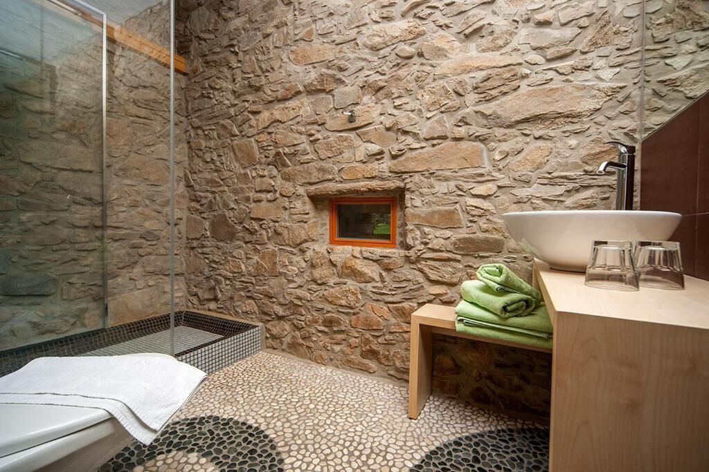 Un baño con pared de piedra, cabina de ducha de vidrio, tocador de madera con un lavamanos grande y una ventana pequeña.