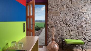 A room with a colorful wall, a stone wall, a wooden door leading outside, a sink, a vase, and a bench.