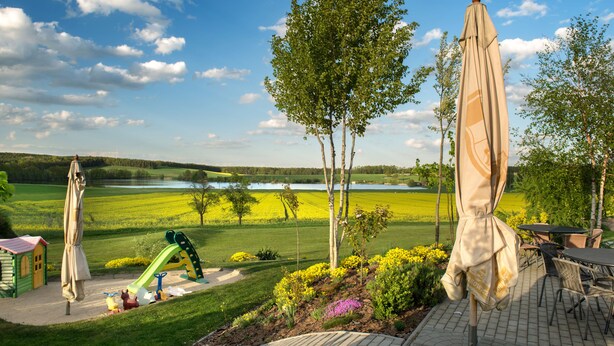A patio area with outdoor seating, a children's play structure, and a view of a lake and green fields. - Penzion Blanský Les (Kvítkovice)