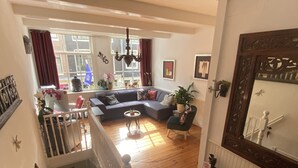A spacious living room with a large sectional sofa, a small round table, and a staircase leading to an upper floor. - Amsterdam B&B Barangay (Amsterdam)
