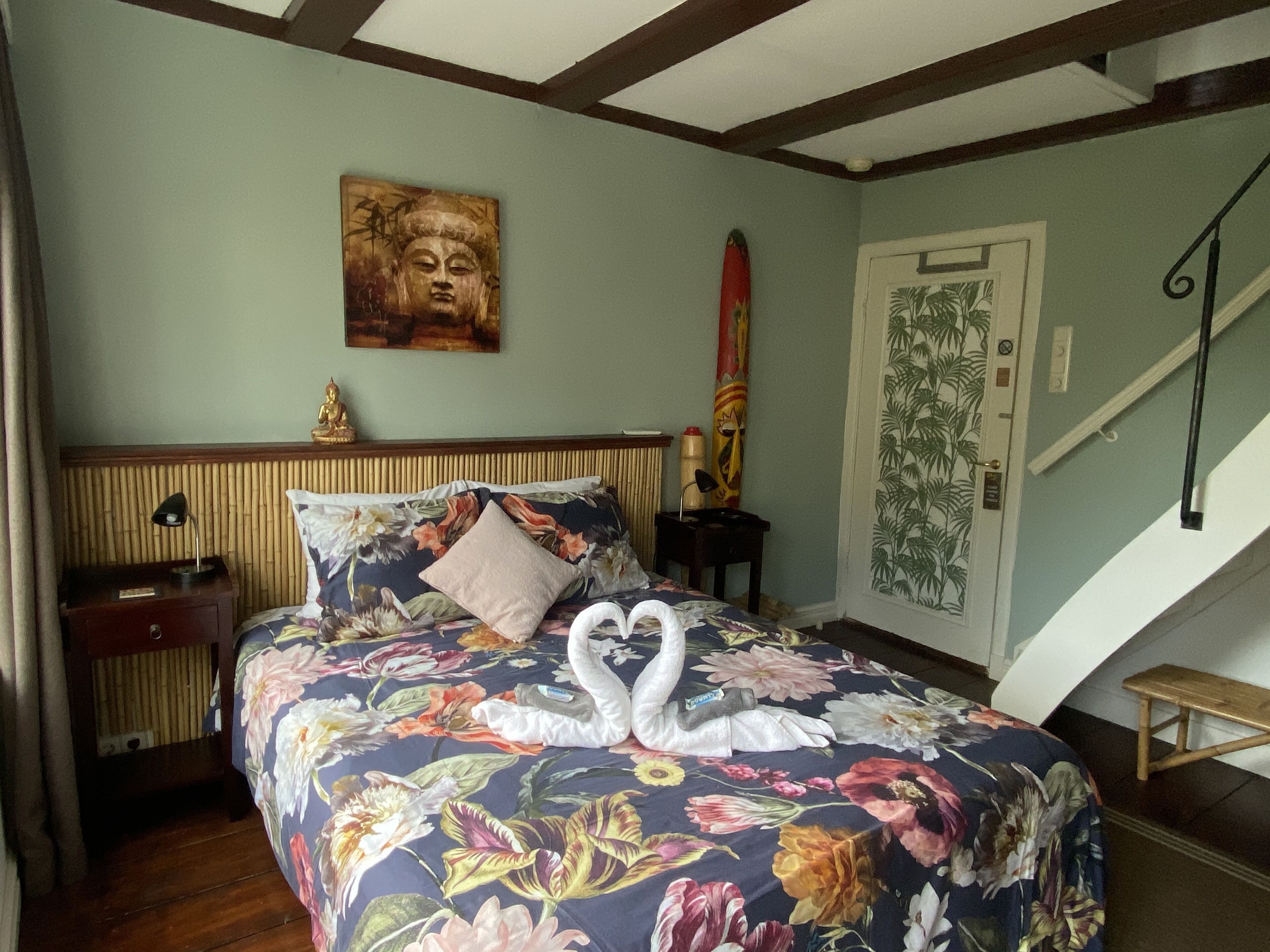 A bedroom with a floral bedspread, a Buddha statue, a swan decoration, a staircase, and a door with a tropical wallpaper design.