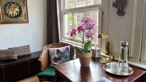 A cozy room with a wooden table, a green chair, a window with a view of buildings, a framed picture on the wall, and a potted orchid on the table. - Amsterdam B&B Barangay (Amsterdam)