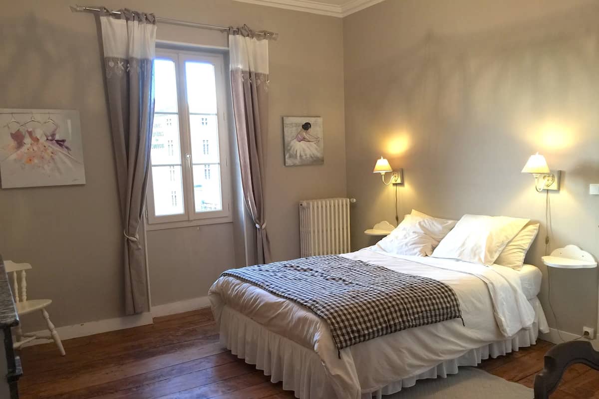 A bedroom with a bed, two bedside lamps, a radiator, a window with curtains, and a painting on the wall.