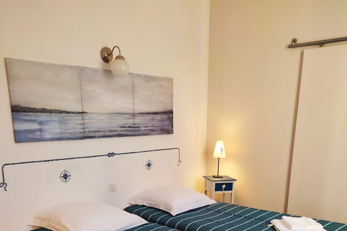A hotel room with two beds, a nightstand with a lamp, a wall-mounted artwork, and a wooden floor.