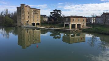 A historic building with a reflection in the water, featuring a stone bridge and a tree in the background.