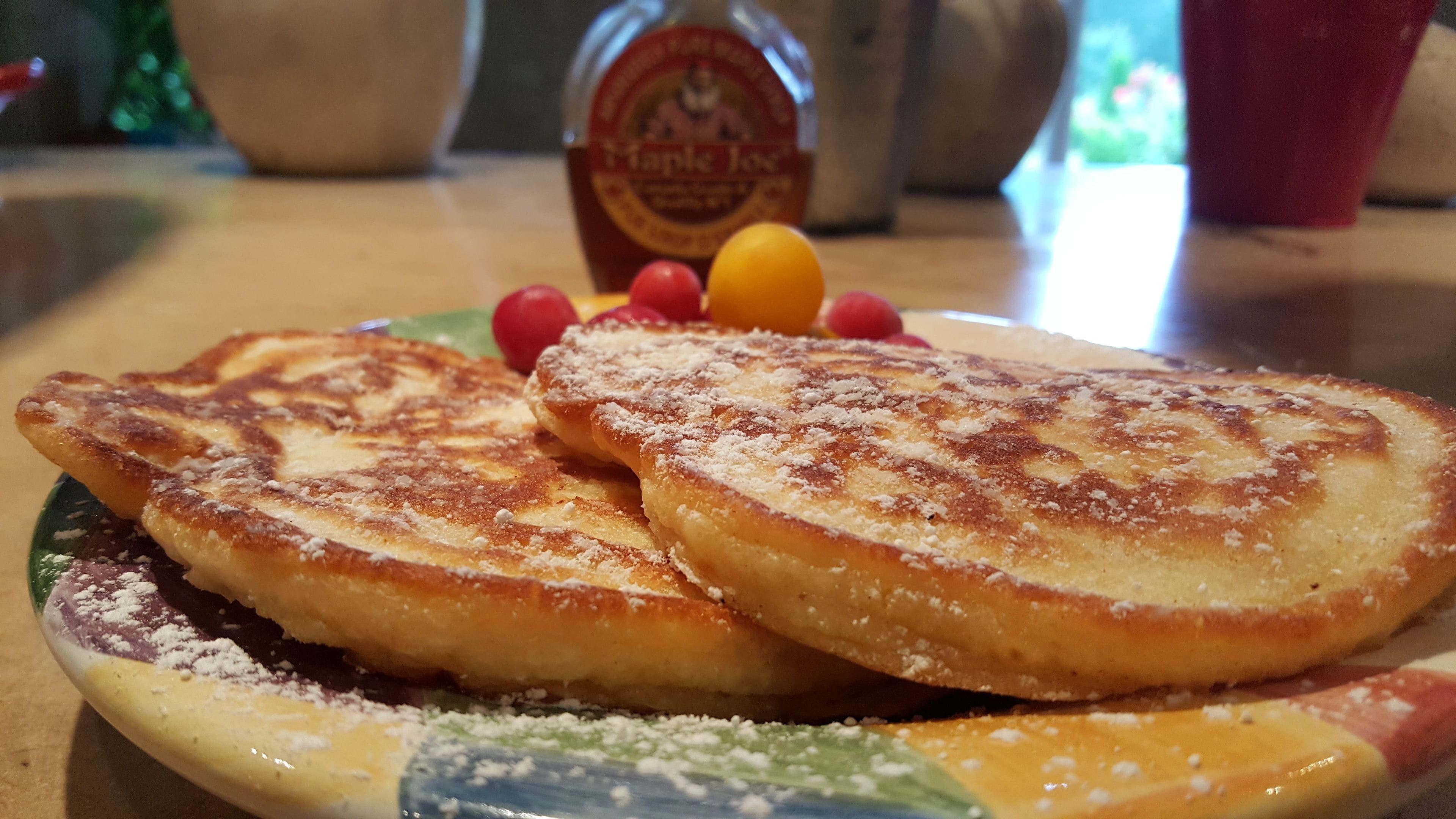 Una porción de tostadas francesas con azúcar glas, acompañada de una botella de sirope de arce y fruta.